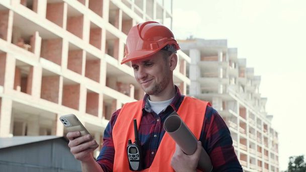Retrato de superintendente de construção sorridente com desenhos e walkie-talkie lendo notícias no celular perto de edifício de vários andares inacabado. Movimento lento - Foto, Imagem