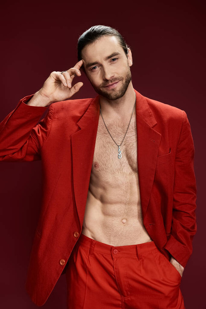 Ein gutaussehender Mann in einem auffallend roten Anzug, ohne Hemd, posiert selbstbewusst in einem Studio-Setting. - Foto, Bild