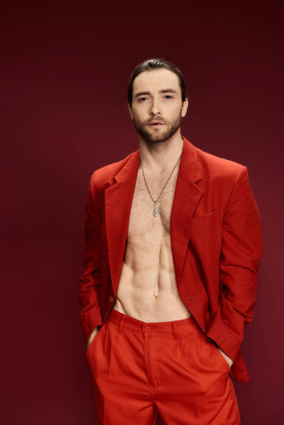 Ein gutaussehender Mann in einem auffallend roten Anzug, ohne Hemd, posiert selbstbewusst in einem Studio-Setting. - Foto, Bild