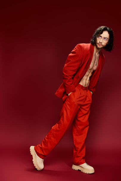 Ένας άντρας χωρίς πουκάμισο αποπνέει αυτοπεποίθηση καθώς ποζάρει για την κάμερα φορώντας ένα εντυπωσιακό κόκκινο κοστούμι, επιδεικνύοντας την τολμηρή και χαρισματική προσωπικότητά του. - Φωτογραφία, εικόνα