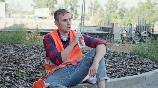 Müder Bauarbeiter sitzt auf einem Bordstein und trinkt Wasser aus einer Plastikflasche, während er in die Ferne blickt. Zeitlupe - Foto, Bild