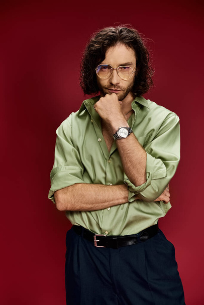 Un hombre guapo con gafas y una camisa de seda verde muestra un reloj de lujo en su mano izquierda sobre un fondo rojo audaz. - Foto, Imagen