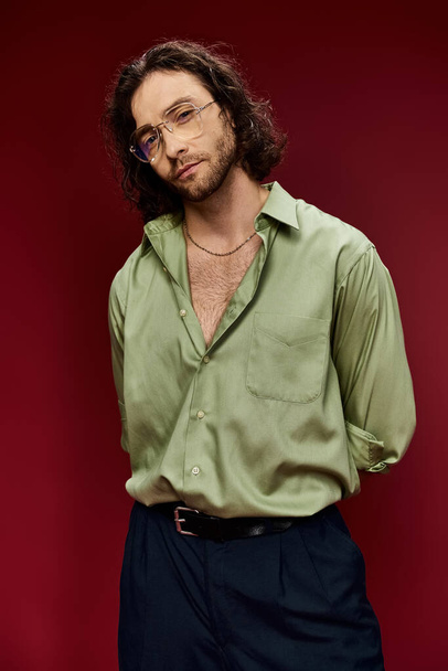 Un homme charismatique aux cheveux longs et aux lunettes portant une chemise de soie verte pose gracieusement sur un fond rouge vif. - Photo, image