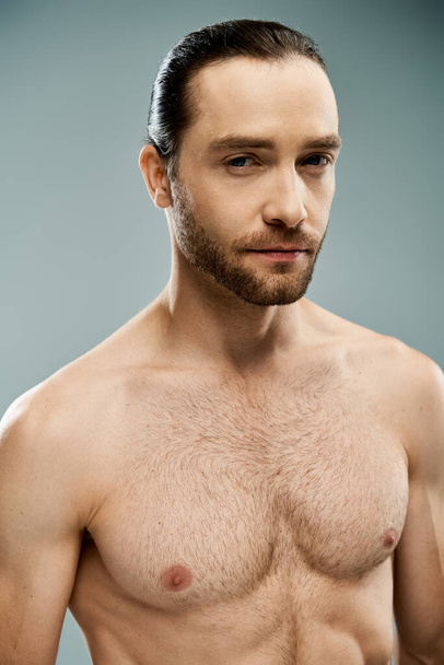 Un hombre guapo con barba se levanta sin camisa sobre un fondo gris en un estudio. - Foto, Imagen