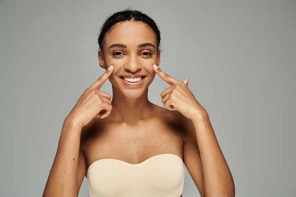Young African American woman in a strapless bra smiling and pointing fingers to the side on a grey background. - Fotografie, Obrázek