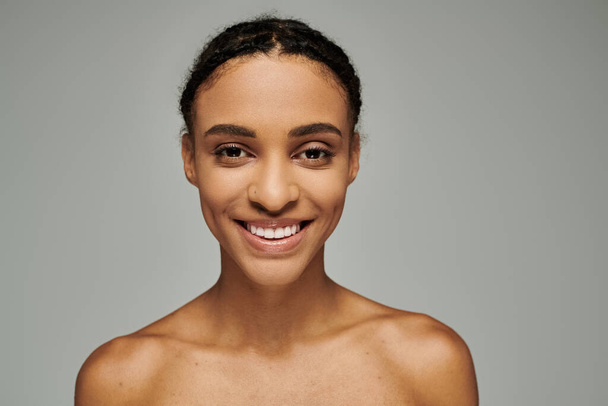 Beautiful young African American woman in a strapless top, exuding joy as she cares for her skin, set against a grey background. - Photo, Image