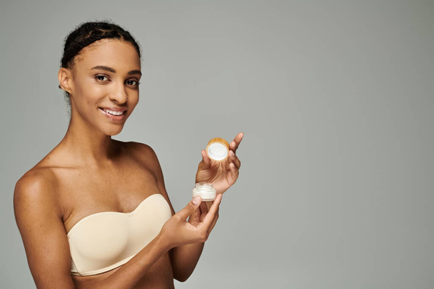 Joven mujer afroamericana en un sujetador cuidando su piel, sosteniendo un recipiente de crema sobre un fondo gris. - Foto, imagen