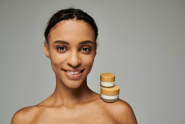 Mujer afroamericana en top sin tirantes sonrisas, frascos de belleza con crema sobre fondo gris. - Foto, imagen