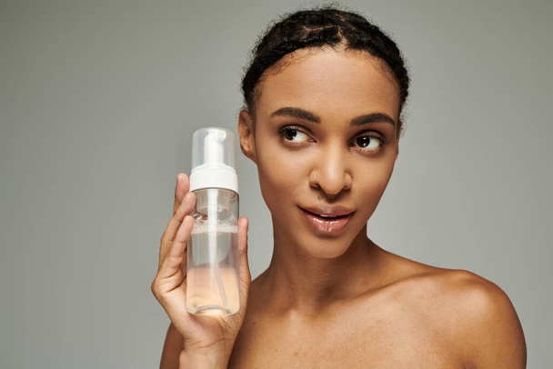 A young African American woman in a strapless top holds a bottle of liquid in front of her face on a grey background. - Foto, Imagem