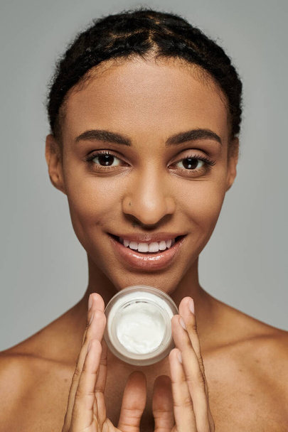 Afro-Amerikaanse vrouw houdt crème pot in de voorkant van gezicht tegen grijze achtergrond, het nemen van huidverzorging serieus. - Foto, afbeelding