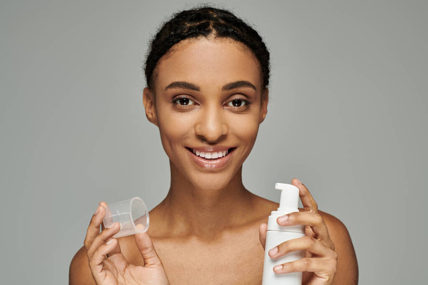 Young African American woman in strapless top holds bottles of cleanser, caring for her skin on a grey background. - Photo, Image