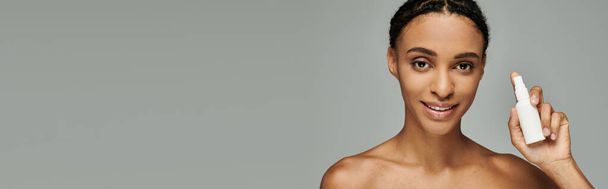 Jonge Afro-Amerikaanse vrouw in strapless top zachtjes houden van een witte huidverzorgingsproduct in haar rechterhand tegen een grijze achtergrond. - Foto, afbeelding