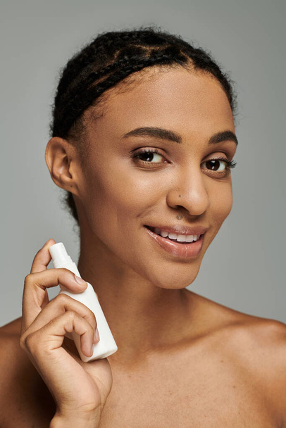 Jonge Afro-Amerikaanse vrouw in strapless top glimlacht terwijl het houden van een cosmetisch product op grijze achtergrond. - Foto, afbeelding