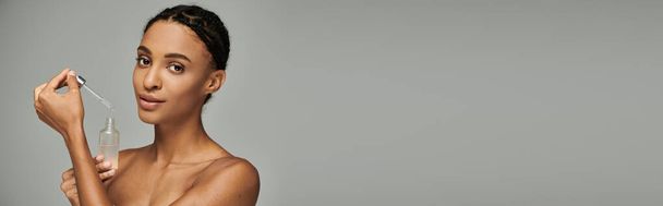 Giovane donna afroamericana in top senza spalline tiene una bottiglia di siero, abbracciando la sua routine di cura della pelle su sfondo grigio. - Foto, immagini