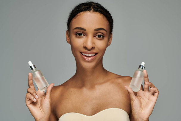 Joven mujer afroamericana en top sin tirantes cuidando la piel, sosteniendo dos botellas de suero sobre fondo gris. - Foto, Imagen
