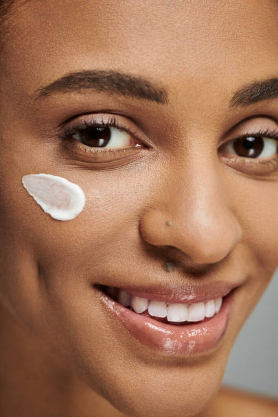 Jeune femme afro-américaine en haut sans bretelles appliquer de la crème sur son visage, en se concentrant sur l'autosoin, routine de soins de la peau. - Photo, image