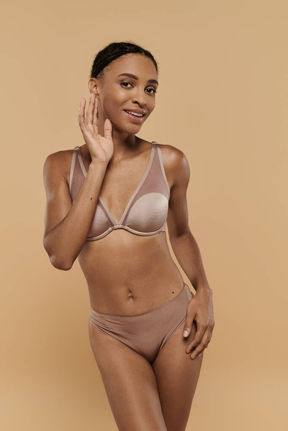 Slim donna afroamericana, indossando un set di lingerie, colpisce una posa fiduciosa su uno sfondo beige. - Foto, immagini