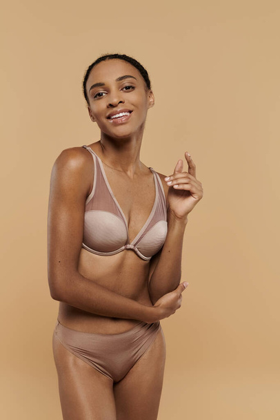 A stunning, slender African American woman elegantly poses in a nude bra, exuding confidence and self-assurance on a soft beige backdrop. - Photo, image