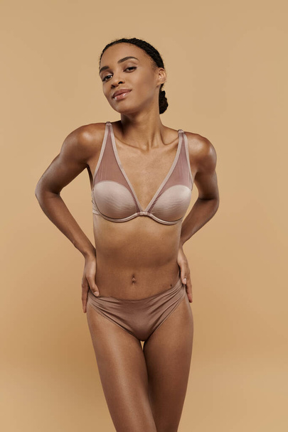 Tyylikäs afroamerikkalainen nainen ruskeissa alusvaatteissa, joka huokuu luottamusta ja huolehtii kehostaan beige-taustalla. - Valokuva, kuva