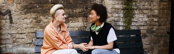 Ein vielfältiges lesbisches Paar sitzt friedlich auf einer Bank in der Nähe einer Ziegelmauer. - Foto, Bild