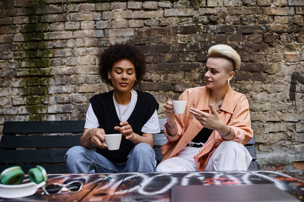 Zwei unterschiedliche Frauen sitzen auf einer Bank und nippen fröhlich an einem Kaffee.. - Foto, Bild