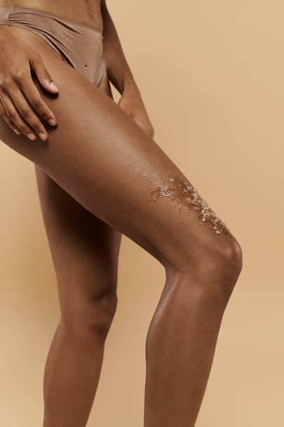 mujer afroamericana recortada con un exfoliante de café en las piernas, cuidando su cuerpo sobre un fondo beige. - Foto, imagen