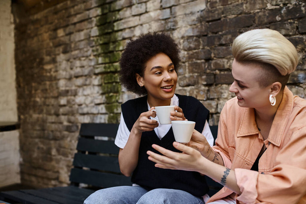 İki farklı lezbiyen kadın bir bankta oturmuş, kahvenin tadını çıkarıyorlar.. - Fotoğraf, Görsel