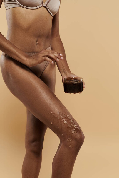 African American woman in bikini with coffee scrub on legs on a beige background. - Photo, Image