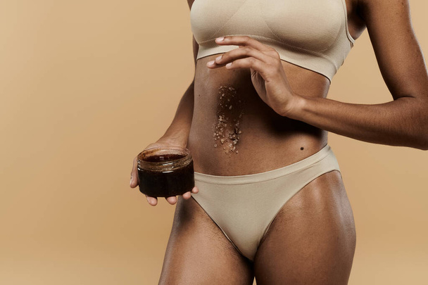 Una splendida donna afroamericana in bikini tiene seducente un barattolo di scrub su uno sfondo beige. - Foto, immagini