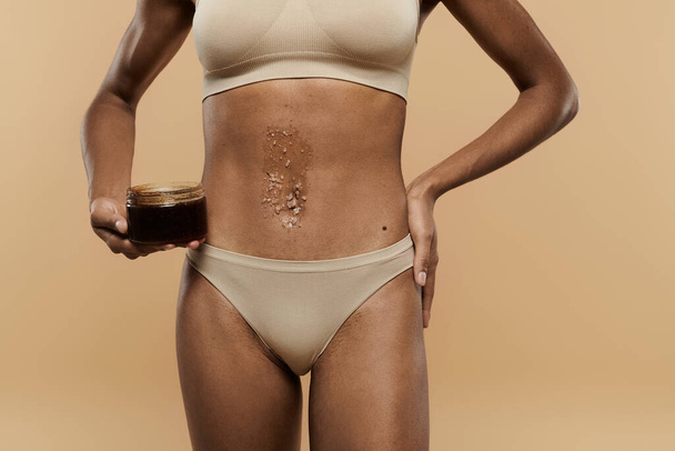 Bikinili, ince Afrikalı Amerikalı bir kadın elinde bir kavanoz çalıyla bej bir arka planda vücuduna bakıyor.. - Fotoğraf, Görsel