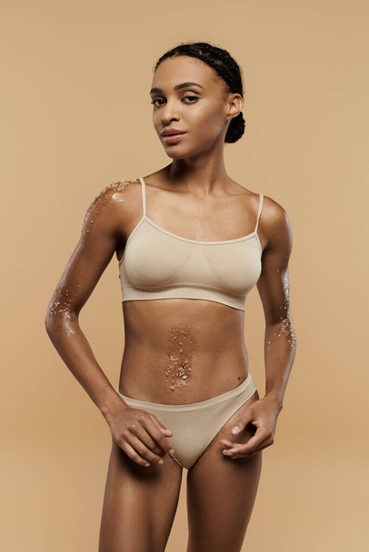 A pretty, slim African American woman is seen taking care of her body in tan lingerie on a beige background. - Photo, Image
