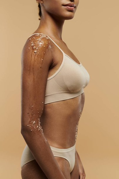 Kaunis ja hoikka afroamerikkalainen nainen huolehtii kehostaan valkoisissa rintaliiveissä ja ruskeissa pikkuhousuissa beige-taustalla.. - Valokuva, kuva