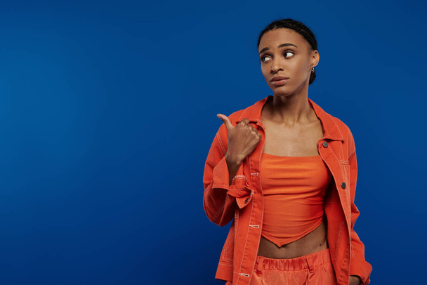 Genç Afrikalı Amerikalı bir kadın mavi arka planda parlak turuncu bir bluz giyerken kendine güvenerek bir şeye işaret ediyor.. - Fotoğraf, Görsel