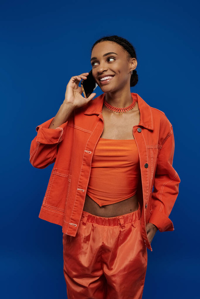 Μια νεαρή Αφρο-Αμερικανίδα με μια ζωηρή πορτοκαλί στολή μιλάει σε ένα κινητό τηλέφωνο σε ένα τολμηρό μπλε φόντο.. - Φωτογραφία, εικόνα