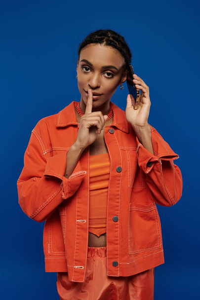 Een mooie jonge Afro-Amerikaanse vrouw die vol vertrouwen in een levendige oranje jas en broek tegen een blauwe achtergrond staat. - Foto, afbeelding