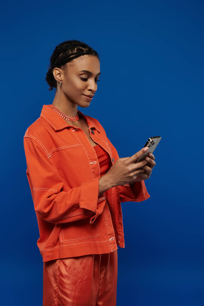 Młoda Afroamerykanka w pomarańczowym stroju pochłonięta telefonem komórkowym na niebieskim tle. - Zdjęcie, obraz