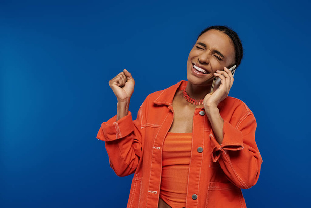 青い背景に対して携帯電話で話しているオレンジ色のシャツの美しい若いアフリカ系アメリカ人女性. - 写真・画像