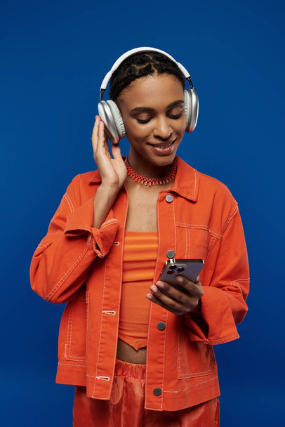 Young African American woman in orange outfit, wearing headphones, engrossed in cellphone screen on blue background. - Photo, Image