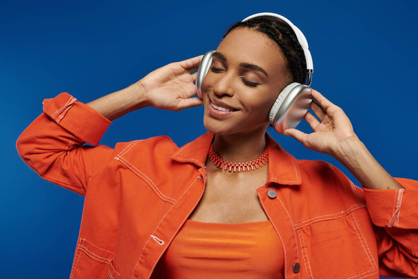 Jonge Afro-Amerikaanse vrouw in oranje shirt genieten van muziek door middel van een koptelefoon op een blauwe achtergrond. - Foto, afbeelding