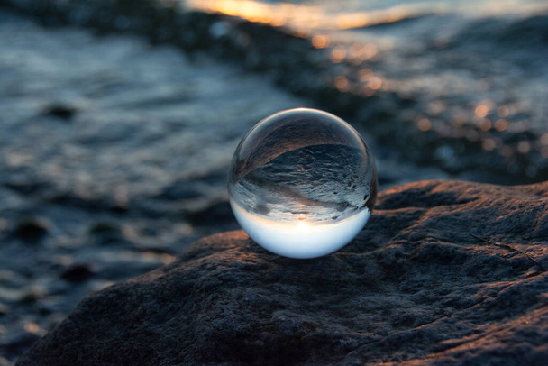 Glaskugel auf einem Felsen bei Sonnenuntergang am Strand, das Meer und die untergehende Sonne spiegeln sich in der Kugel - Foto, Bild