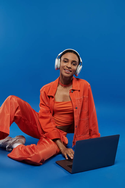 Żywa młoda Afroamerykanka w pomarańczowym stroju siedzi na podłodze, pochłonięta laptopem ze słuchawkami. - Zdjęcie, obraz