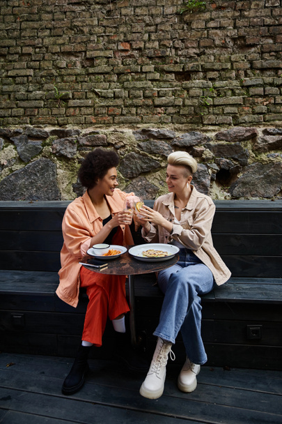 Dwie kobiety, różnorodna lesbijska para, jedzą razem posiłek na ławce w kawiarni. - Zdjęcie, obraz