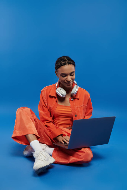 Egy fiatal afro-amerikai nő élénk narancssárga ruhában ül a padlón, és egy laptopon gépel, kék háttérrel.. - Fotó, kép