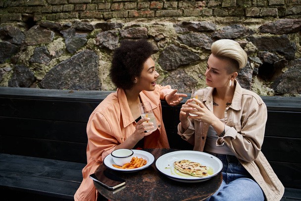 Ένα ποικιλόμορφο ζευγάρι λεσβιών κάθεται σε ένα τραπέζι με πιάτα νόστιμο φαγητό σε ένα καφέ. - Φωτογραφία, εικόνα