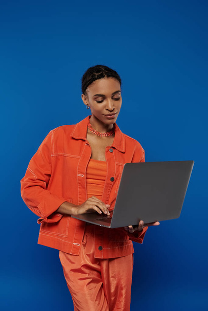 Una giovane donna afroamericana in tuta arancione con un computer portatile sullo sfondo blu. - Foto, immagini