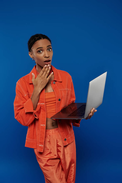 Nuori afrikkalainen amerikkalainen nainen oranssissa asussa pitää luottavaisesti kannettavaa tietokonetta elinvoimaista sinistä taustaa vasten. - Valokuva, kuva