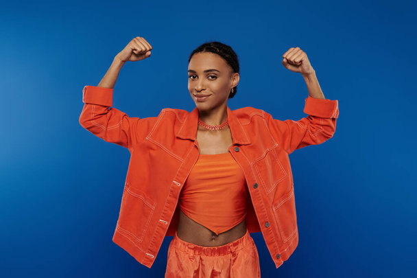 Egy csinos fiatal afro-amerikai nő kiemelkedik élénk narancssárga dzsekiben és rövidnadrágban, kék háttérrel.. - Fotó, kép