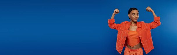 Nuori afroamerikkalainen nainen seisoo luottavaisesti eloisassa oranssissa paidassa ja housuissa sinistä taustaa vasten. - Valokuva, kuva