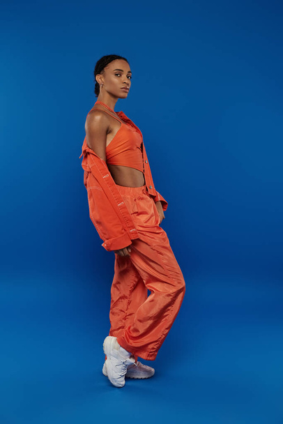 Досить молода афроамериканка в яскравому помаранчевому вбранні позує на вражаючий синій фон. - Фото, зображення