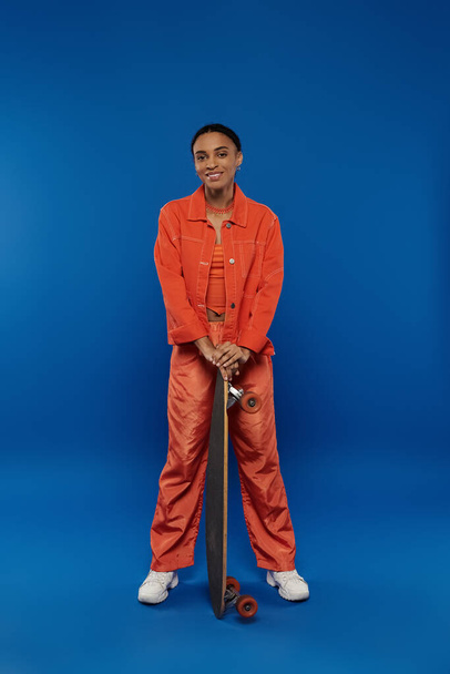A young African American woman in an orange jumpsuit holding a skateboard on a vibrant blue background. - Photo, Image